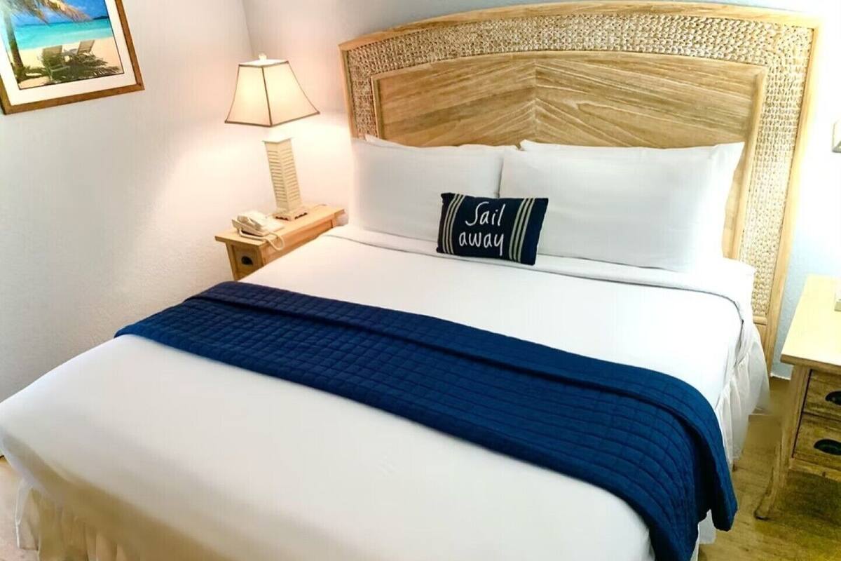 A neatly made bed with a blue blanket, a pillow with "Jail away" text, a bedside lamp, and a small wooden nightstand with a phone.