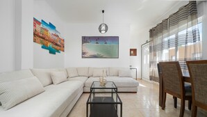 A living room with a sectional sofa, a glass coffee table, a dining area with chairs, a painting, and a window with curtains. - Holiday Home 'Linda' with Private Terrace, Wi-Fi and Air Conditioning (Olhão)