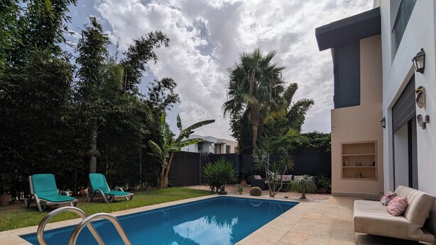 Una piscina con una escalera metálica, rodeada por un patio con suelo de baldosas. Hay sillones de descanso y un sofá con cojines.