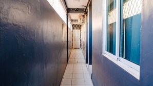 Un couloir étroit au sol carrelé, avec un mur bleu d’un côté et une fenêtre de l’autre.
