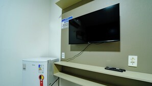 Une chambre dotée d’un téléviseur à écran plat fixé au mur, d’un petit réfrigérateur et d’une étagère sur laquelle se trouve une télécommande.