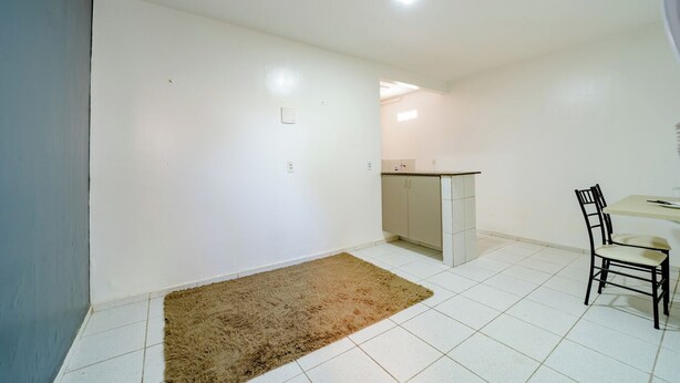 Une petite pièce propre aux murs blancs, dotée d’un sol carrelé, d’un tapis marron, d’une kitchenette et d’un coin repas avec une chaise et une table.