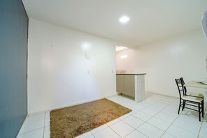 A small, clean room with white walls, a tiled floor, a brown rug, a kitchenette, and a dining area with a chair and table. - Flat c Wi-fi e Ar-condicionado Perto do Aeroporto (Fortaleza)