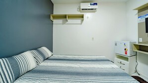 Une petite pièce, peu meublée, comprenant un lit, un climatiseur fixé au mur et un petit réfrigérateur blanc.