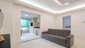A modern living room with a grey sofa, a flat-screen TV, and a kitchen area visible through an open doorway. - Apt w/ Balcony & BBQ 290m From the sea Cau0199 (Itapema)