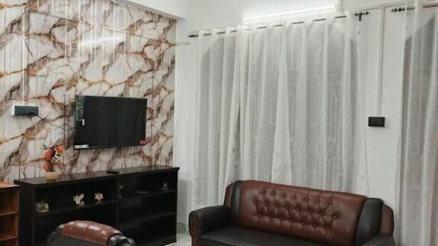 A living room with a marble-patterned wall, a leather sofa, two armchairs, a glass coffee table, a ceiling fan, and a TV mounted on the wall. - Rudra homes (Guwahati)