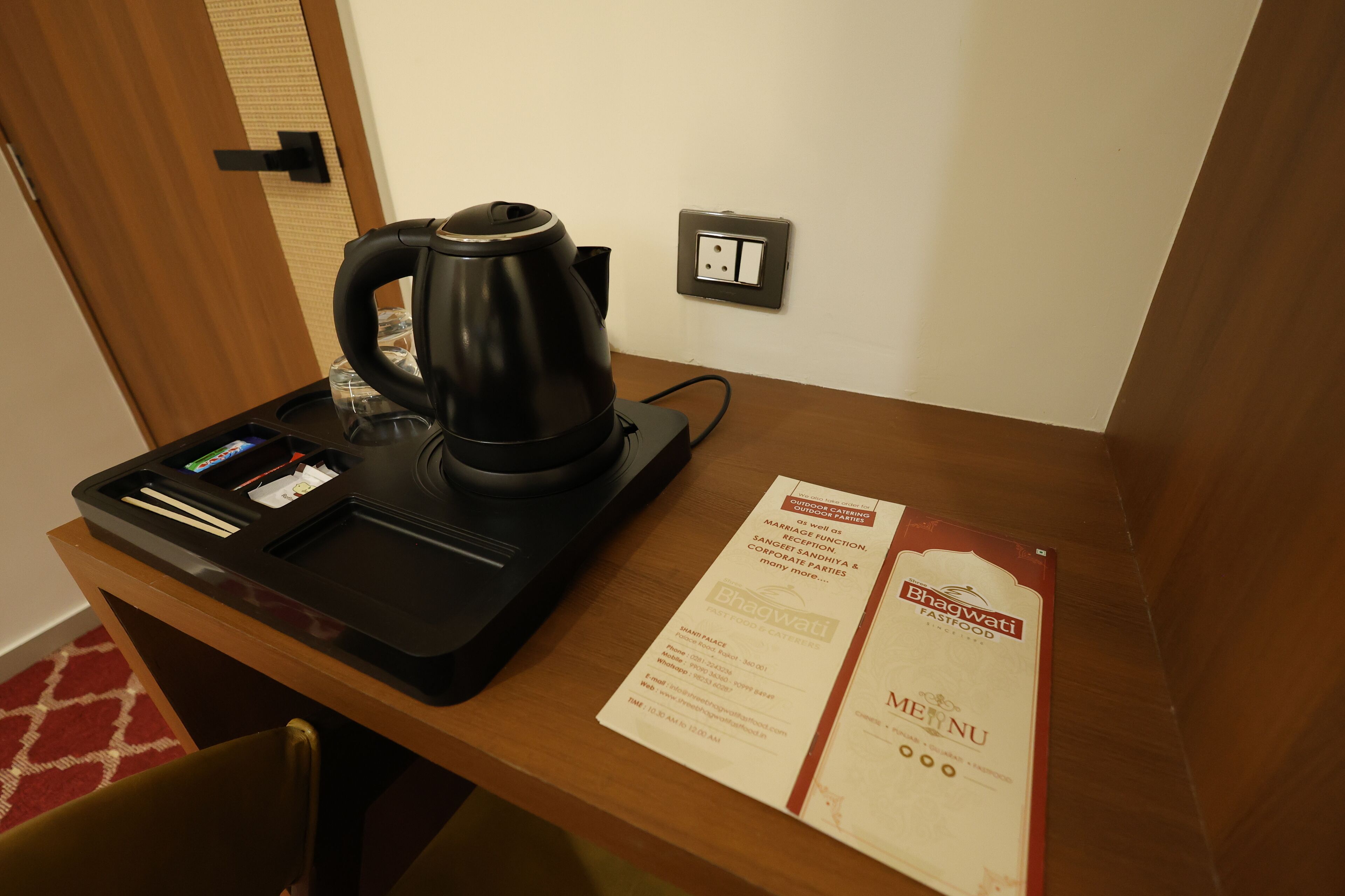 A hotel room with a wooden desk, a black electric kettle, a tray with a smartphone and a glass, and a menu on the wall.