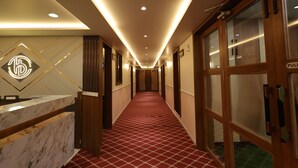 A hotel reception area with a marble counter, a red patterned carpet, and a long hallway with doors and windows. - Hotel Heaven Blue (Rajkot)
