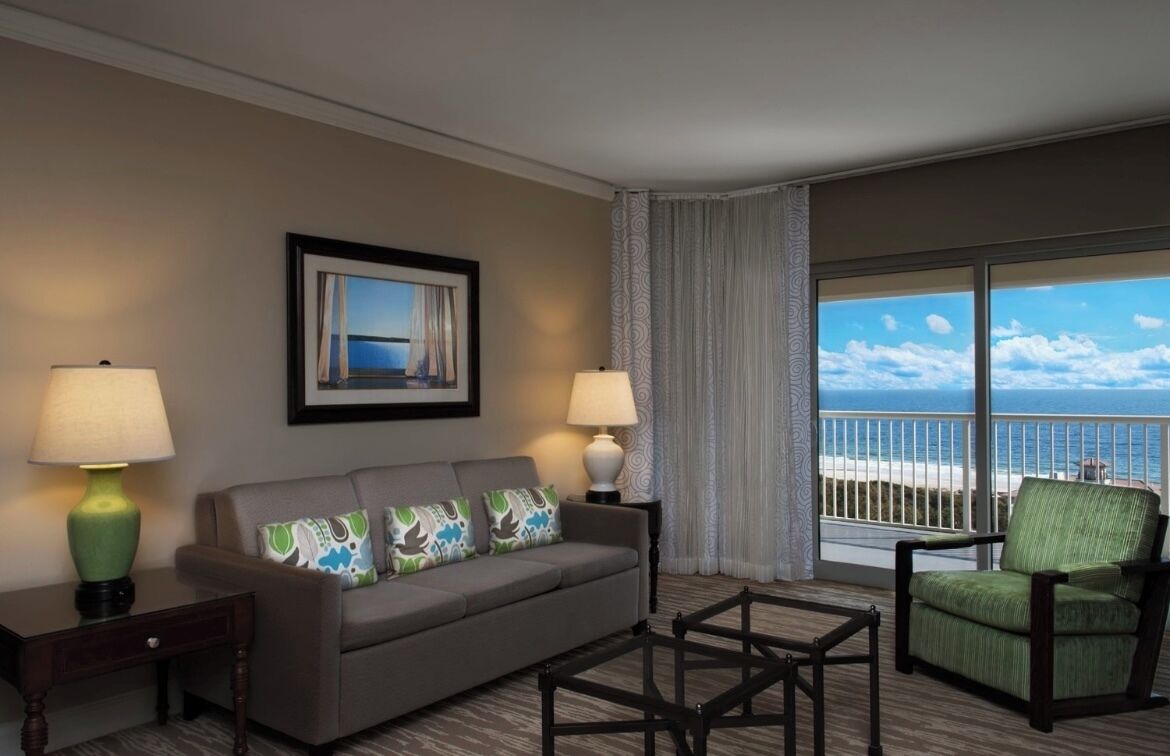A hotel room with a balcony, a sofa, a chair, and two lamps.