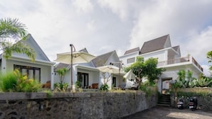 A row of white houses with sloped roofs, a stone wall, and a paved area with parked scooters. - Villa Dangka Kintamani Mitra RedDoorz (Bali)