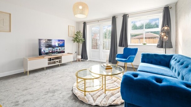 Ett modernt vardagsrum med en blå soffa, ett soffbord i glas, en tv på ett tv-bord och ett stort fönster med gardiner.