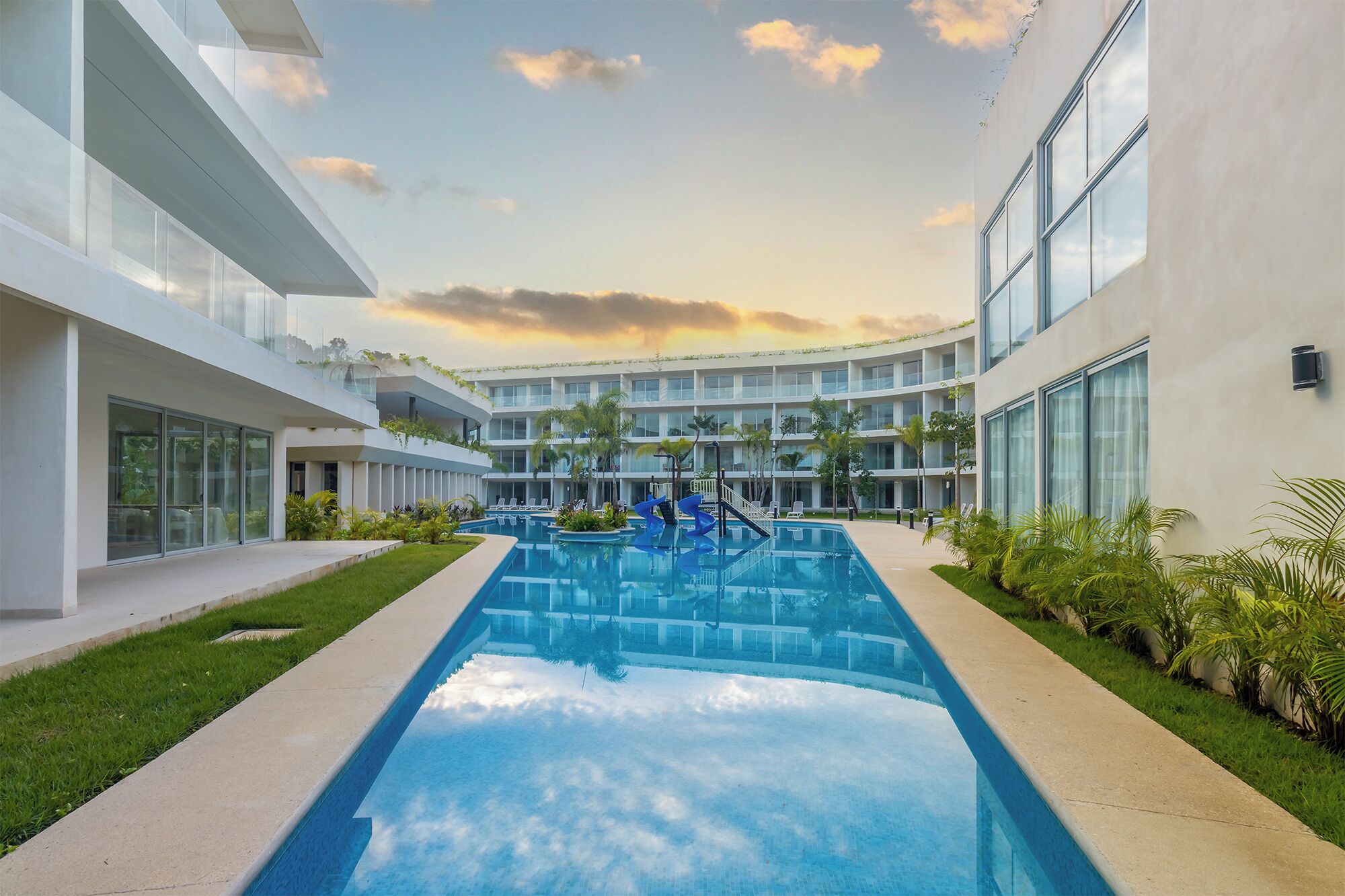 A modern hotel with a central swimming pool, multiple balconies, and lush greenery.