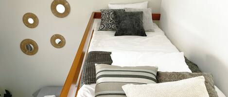 A single bed with a wooden frame, white bedding, and multiple pillows in various patterns and textures.