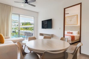 Habitación de hotel con una mesa redonda, sillas, cama con cabecera, televisor montado en la pared y ventana con vista a una piscina y edificios.