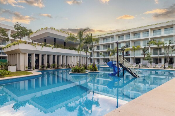 Un hotel moderno con piscina central, varios balcones y exuberante vegetación.
