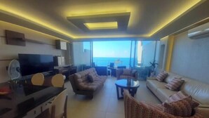A modern living room with a flat-screen TV, a sofa, a patterned armchair, and a dining area with a table and chairs.