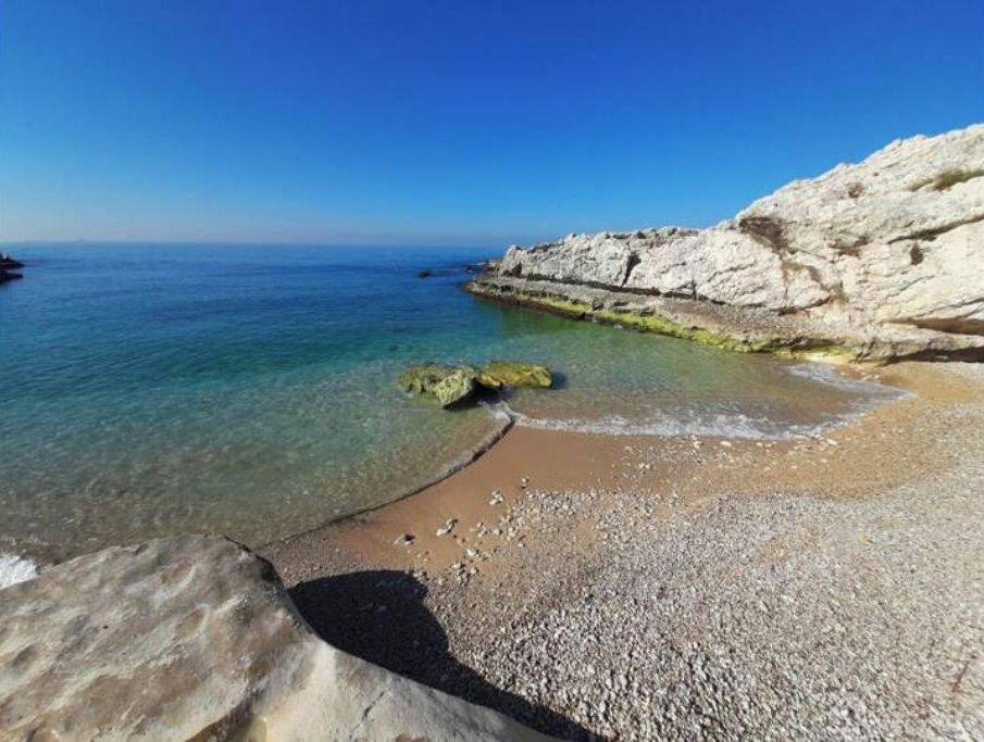 Une plage côtière à l’eau claire, avec un rivage de galets et des formations rocheuses blanches.