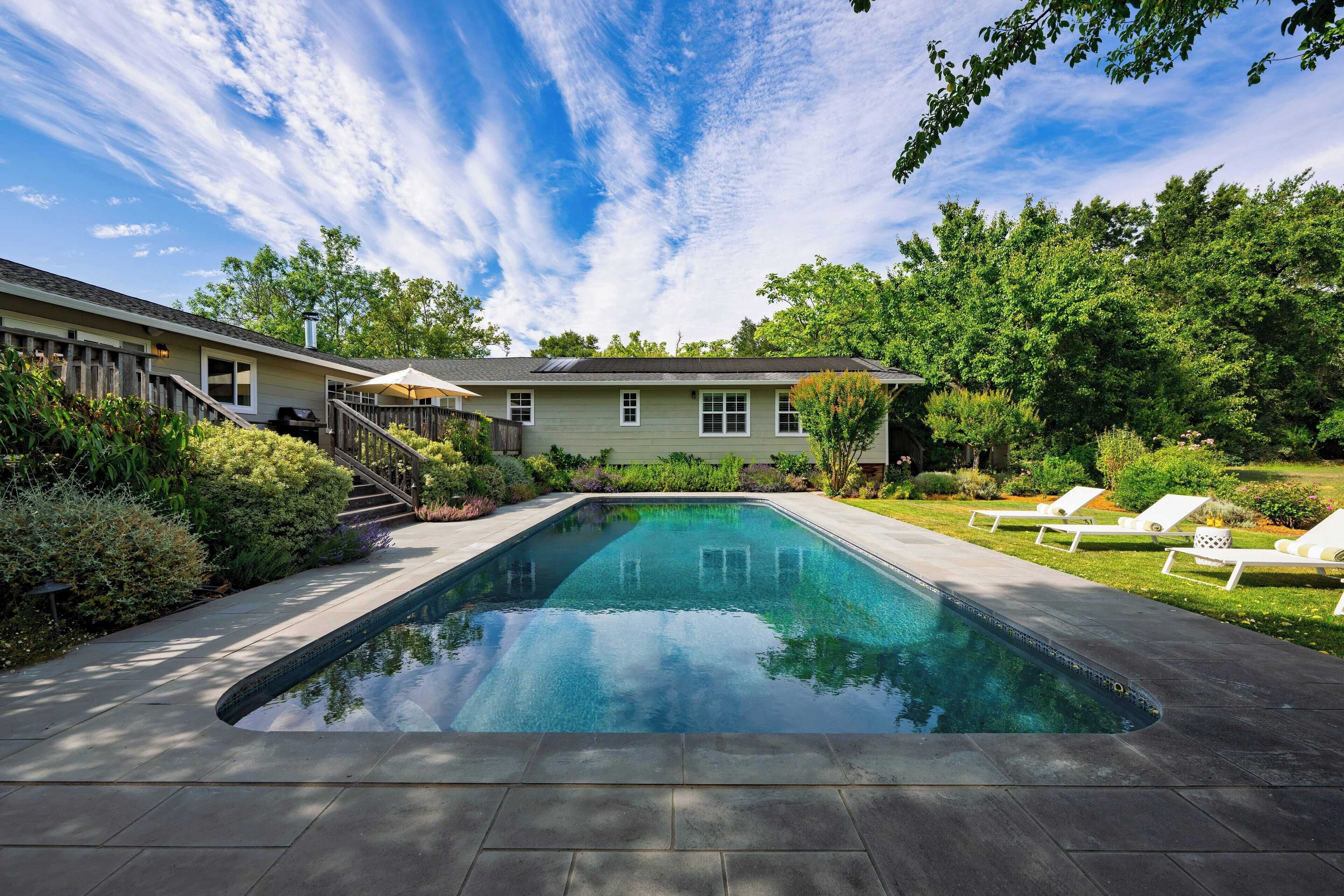 A swimming pool with a wooden deck, surrounded by lush greenery and a house with a covered patio.