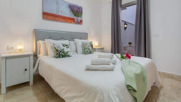 A neatly made bed with white linens and a green throw, a nightstand with a lamp, and a painting above the bed. - Holiday Apartment 'Rolycar 101' with Private Terrace and Wi-Fi (Icod de los Vinos)