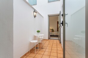 Un pasillo estrecho con piso de baldosas, una pequeña mesa con una planta en maceta, y una puerta que da a una habitación con una cama y un edredón estampado.