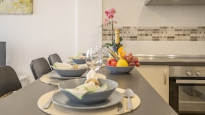 Une cuisine moderne avec une table à manger dressée pour deux personnes, un panier de fruits, une bouteille de champagne et une orchidée décorative.