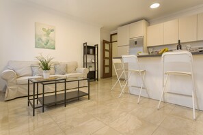 A living room with a sofa, a coffee table, and a plant. There is a kitchen area with a refrigerator, a countertop, and a bar stool.