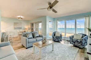 A spacious living room with a view of the ocean, a ceiling fan, and a light-colored rug.