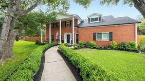 A two-story brick house with a covered front porch, a flag, and a well-maintained lawn. - Austin's Hill Country Escape & Retreat – Private Oasis With Pool & Spa on Acre+ (Austin)