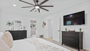 A bedroom with a bed, two dressers, a TV, a ceiling fan, and a door leading to another room. - Austin's Hill Country Escape & Retreat – Private Oasis With Pool & Spa on Acre+ (Austin)