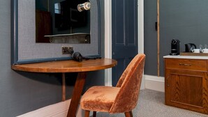 A room with a round wooden table, a velvet chair, a mirror, a door, and a wooden cabinet with a coffee maker and glasses.