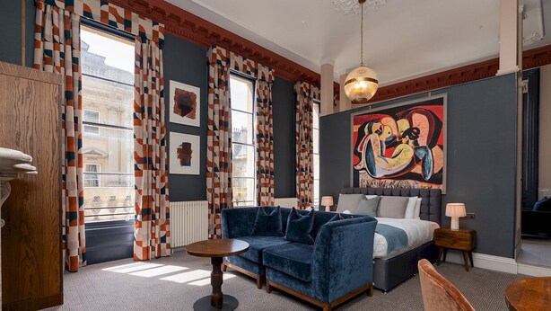 A modern hotel room with a blue sofa, a bed, a round table, and a large painting on the wall.