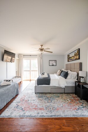 A bedroom with a bed, a ceiling fan, a bedside table with a lamp, a TV mounted on the wall, and a window with curtains.