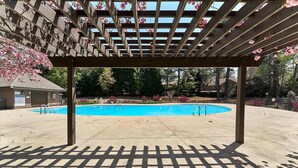 Une piscine extérieure couverte, surmontée d’une pergola en bois, avec un ciel bleu dégagé.