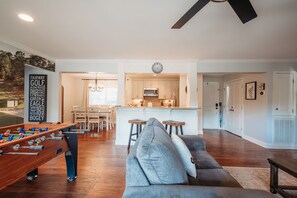 En moderne stue med et fodboldbord, en grå sofa og et køkkenområde med træskabe og en central ø.