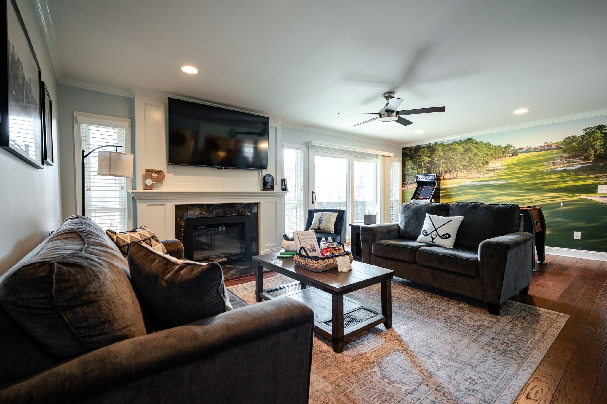 A living room with a fireplace, a television mounted above it, a ceiling fan, and a large window with a view of a golf course.