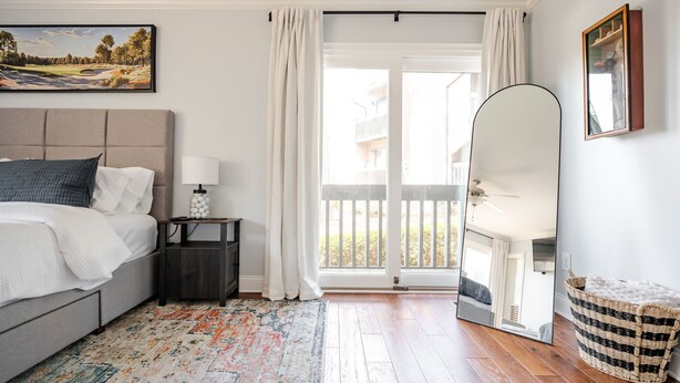 Une chambre à coucher comprenant un lit, une table de chevet, une lampe, un grand miroir, un panier en osier et une vue sur l’extérieur grâce à une porte coulissante.