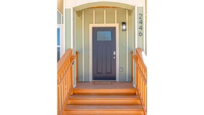 Una escalera de madera conduce a una puerta oscura con un ventanuilla pequeña, flanqueada por paredes de tonos claros y un buzón.