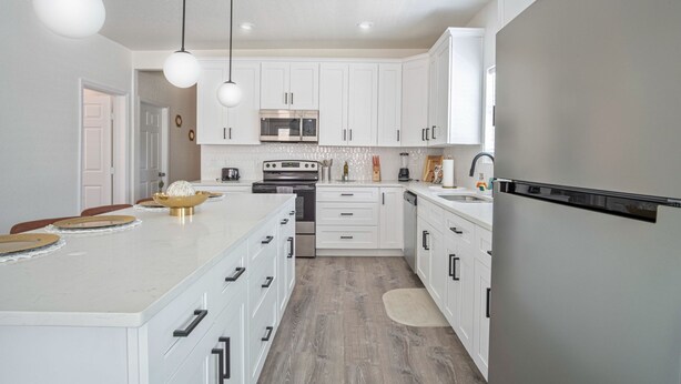 Una cocina moderna con armarios blancos, una isla grande, electrodomésticos de acero inoxidable e iluminación colgante.