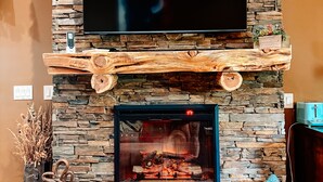 A stone fireplace with a flat-screen TV above it, a wooden mantel, and a small shelf with a vase of dried plants. - Bison Suite - relax in your private hot tub and explore the Ranch!  (Valley View)