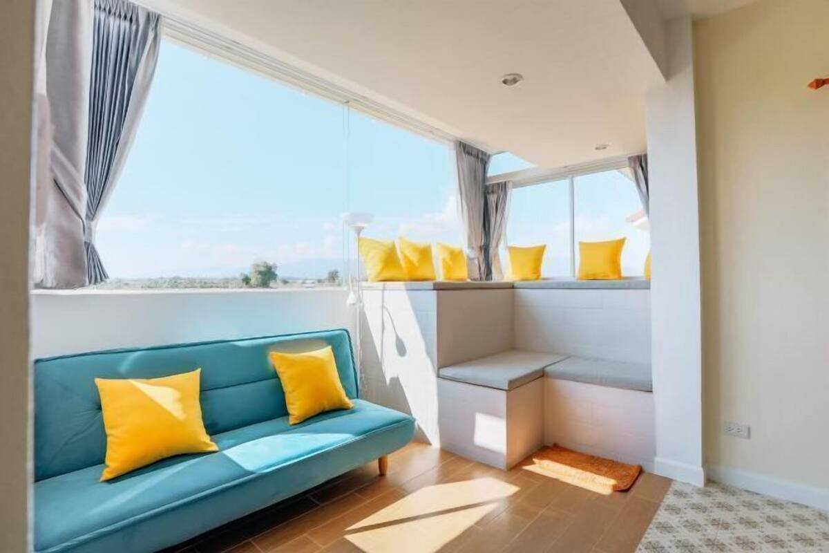 A modern room with a blue sofa, yellow pillows, a small table, and large windows offering a view.