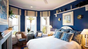 A bedroom with a large bed, two chairs, a desk, a fireplace, and a view of palm trees. - Snug Harbor Inn (Avalon)