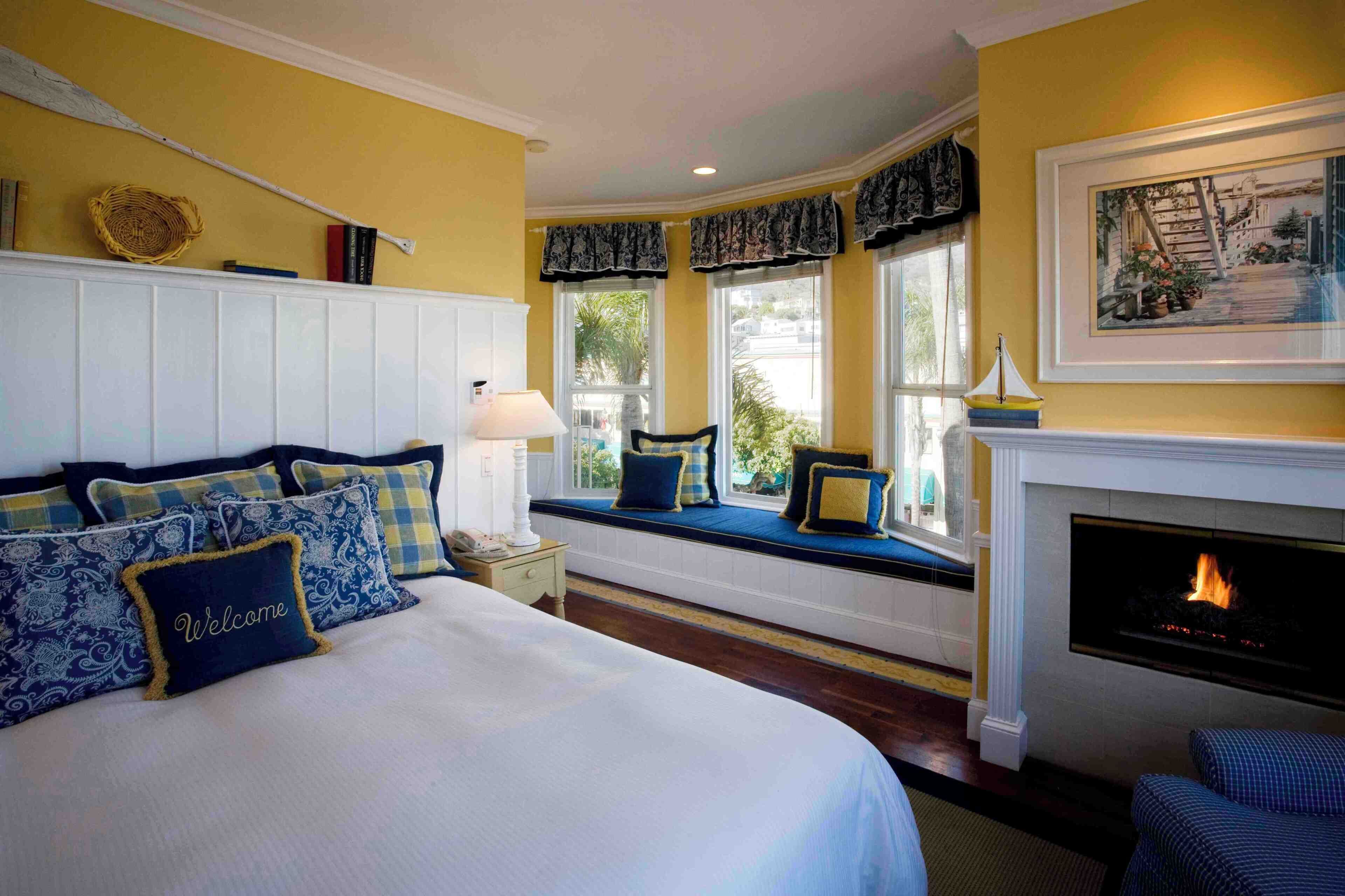 A bedroom with a bed, a fireplace, a bay window with seating, and a view of the outdoors.