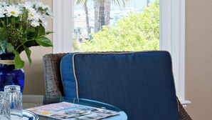 A room with a large window, a wicker chair, a table with a vase of flowers, and a magazine. - Snug Harbor Inn (Avalon)