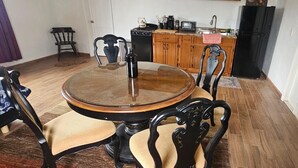 Uma sala de jantar com uma mesa redonda, quatro cadeiras, uma cozinha ao fundo e uma porta  à direita.