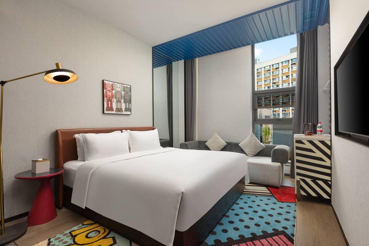 A hotel room with a bed, a desk, a chair, a sofa, a TV, and a window with a view of buildings.