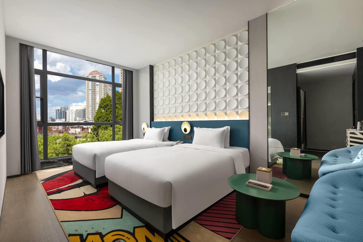 A modern hotel room with two beds, a large window offering a city view, a unique wall design, and a small seating area.