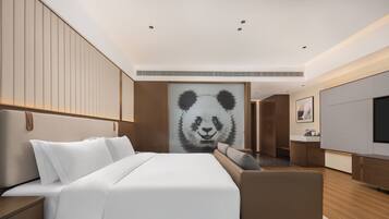 A modern hotel room with a large bed, a sofa, a TV, and a panda-themed artwork on the wall.