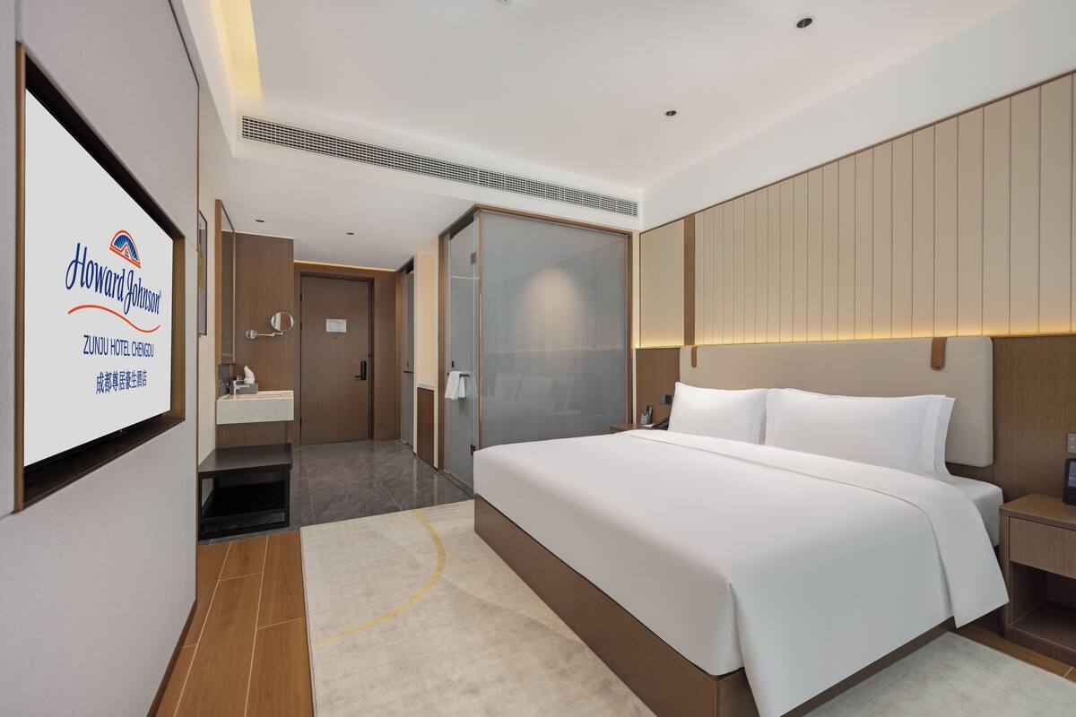 Ein modernes Hotelzimmer mit einem großen Bett, einem Nachttisch, einer Duschkabine aus Glas, einem Schminktisch mit Spiegel und einem an der Wand befestigten Fernseher, der den Namen des Hotels anzeigt.