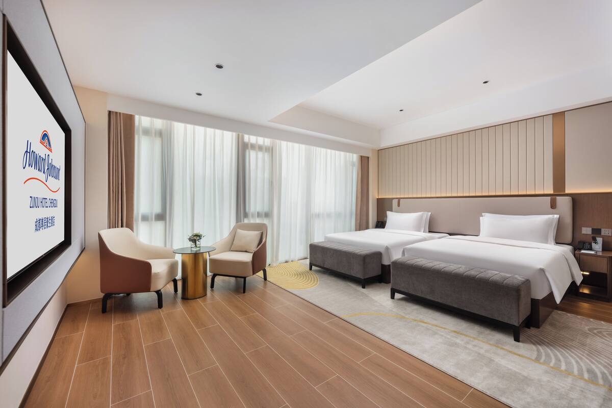 Ein Hotelzimmer mit zwei Betten, einem Schlafsofa, einem kleinen Tisch und einem Fernsehbildschirm, der die Hotelmarke zeigt.
