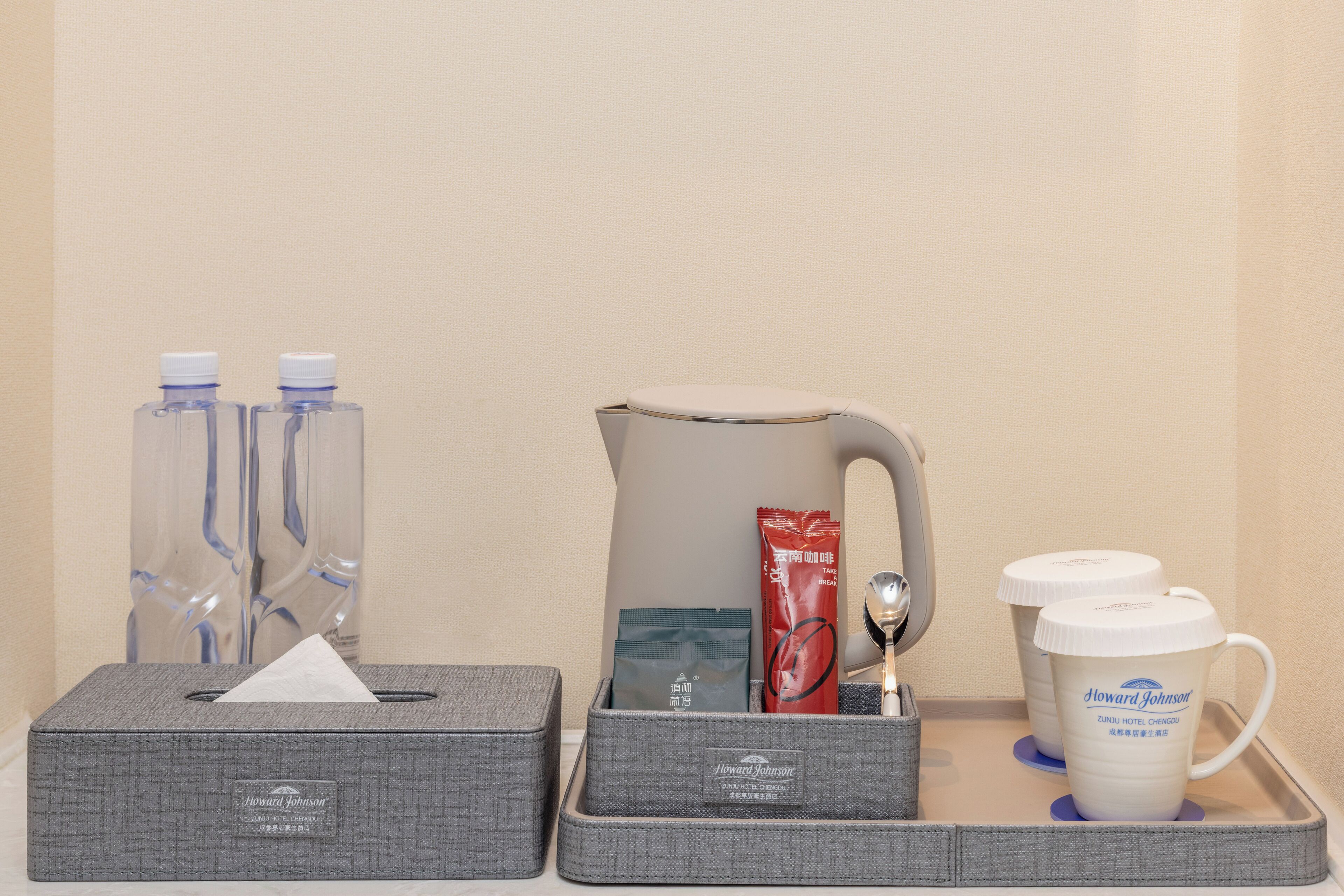 A tray with a tissue box, water bottles, a coffee pot, a packet of tea, a spoon, and two coffee mugs.